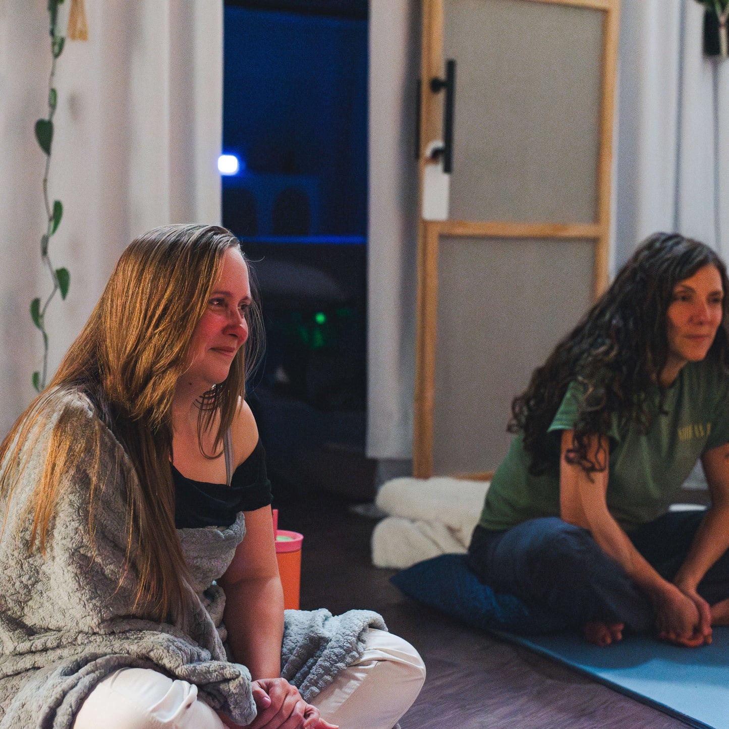 Two women sitting on yoga mats in a room with curtains and a door.