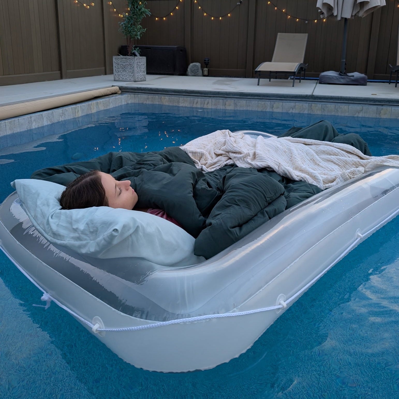 Person lying on an inflatable pool bed in a swimming pool with a fence and plants in the background.