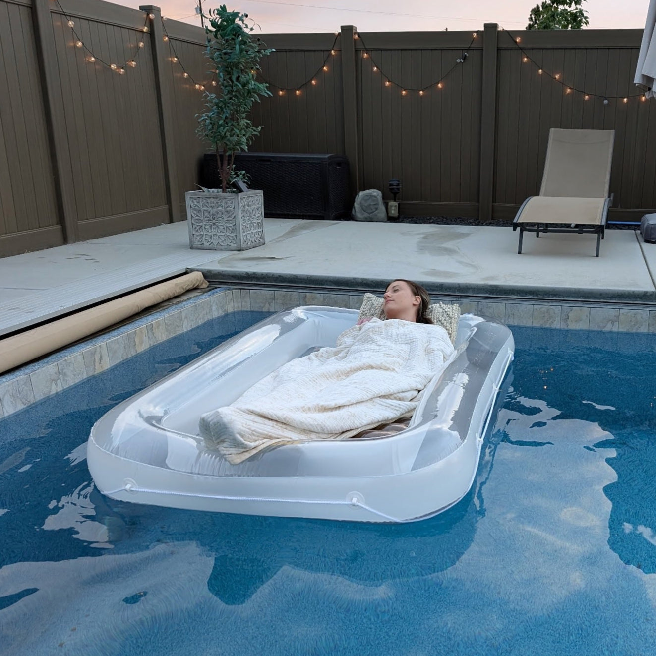 Person lying on a pool float in a backyard pool with string lights and chairs.