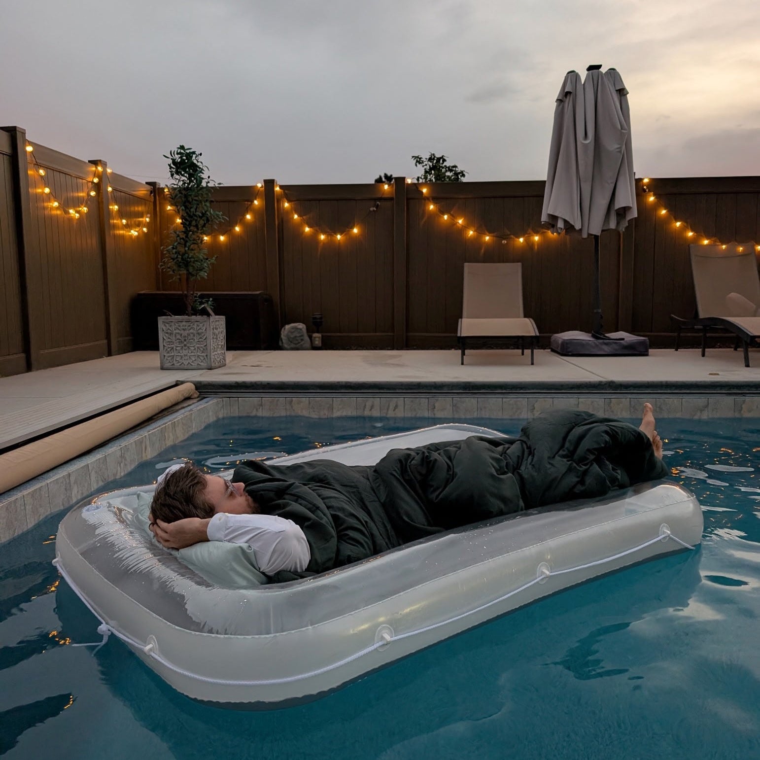 Person lying on an inflatable pool raft in a backyard pool with string lights and outdoor furniture.